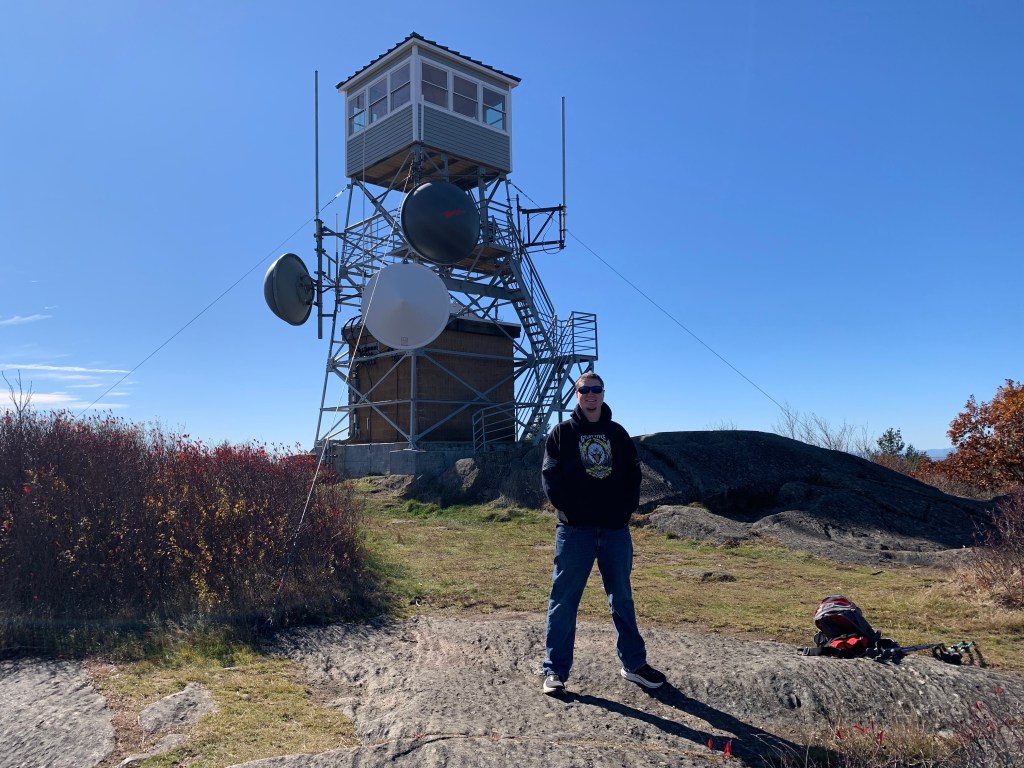 My grown son standing in front of the fire tower at the top of Pitcher Mountain in New Hampshire.