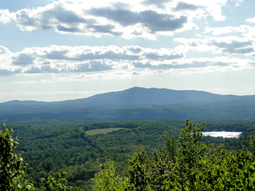 A panoramic view of a mountainous landscape with lush greenery in the foreground and a serene lake visible among the trees, under a partly cloudy sky.