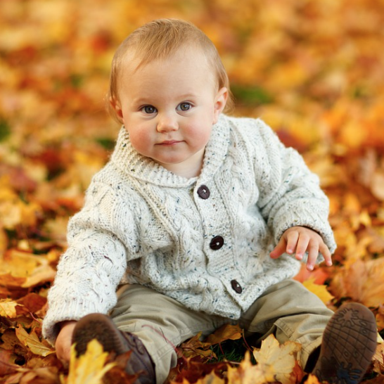 Baby in knitted, button up cardigan sitting in fall leaves.