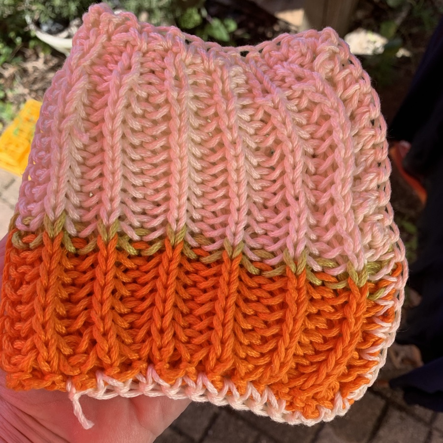 A hand holding a handmade crochet piece in shades of pink, orange, and green, featuring a textured pattern.