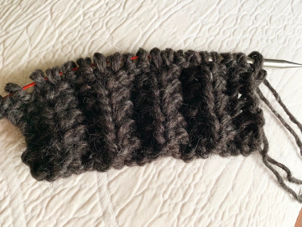 Close-up of a partially knitted dark gray yarn project on knitting needles, resting on a textured white surface.