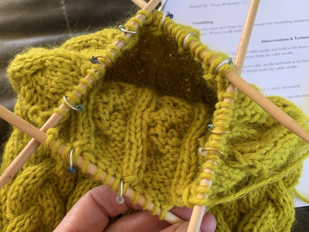 Close-up of a person's hand holding yellow knitted fabric with wooden knitting needles, showing progress on a project, with a printed pattern in the background.