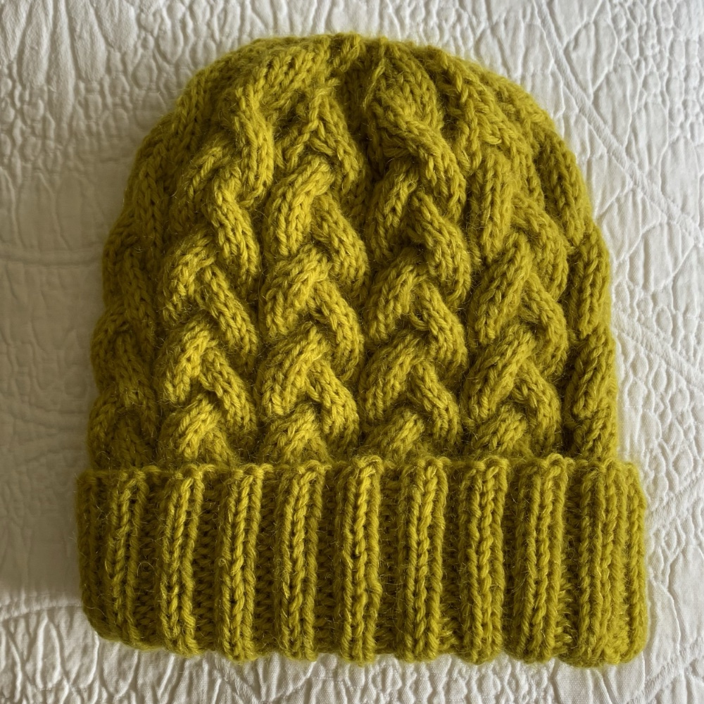 Bright yellow knitted beanie hat with a cable pattern, displayed on a textured white surface.