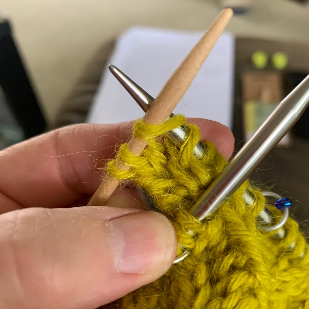 Close-up of a hand holding knitting needles with yellow yarn, focusing on the stitches being worked.